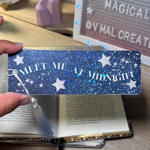 May include: A navy blue glitter bookmark with the text "MEET ME AT MIDNIGHT" in white, along with white star accents. The bookmark has a silver tassel attached. The bookmark is placed in an open book.