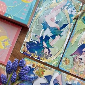 Puede incluir: Una colecci&oacute;n de tarjetas rectangulares con ilustraciones estilo anime de personajes con trajes azules, verdes y amarillos con bordes de filigrana dorada. Las tarjetas muestran motivos celestiales, estrellas y lunas, junto a flores moradas.