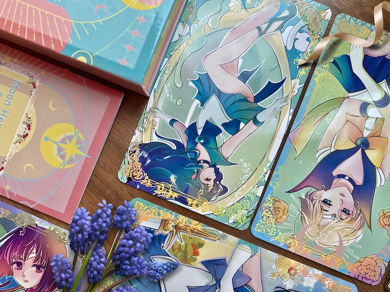 Puede incluir: Una colecci&oacute;n de tarjetas rectangulares con ilustraciones estilo anime de personajes con trajes azules, verdes y amarillos con bordes de filigrana dorada. Las tarjetas muestran motivos celestiales, estrellas y lunas, junto a flores moradas.