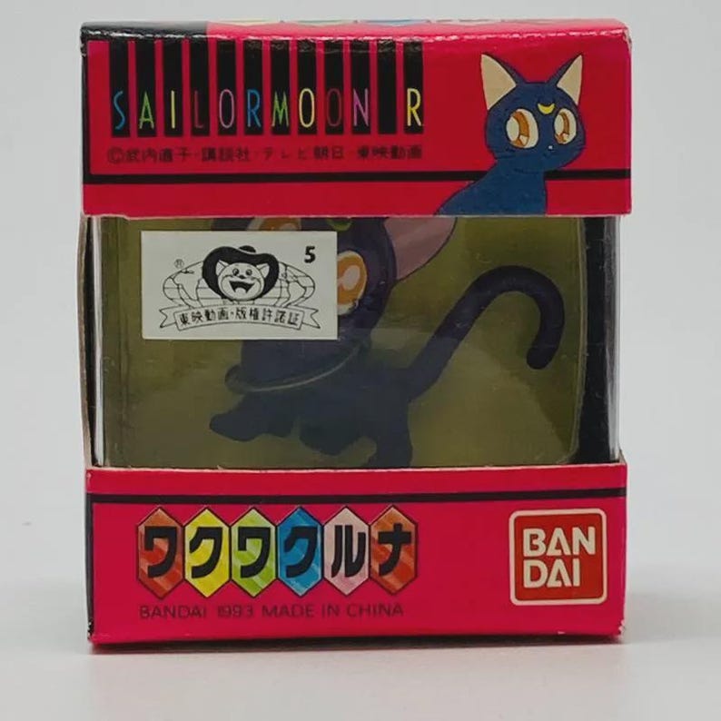 Op de afbeelding: Een vintage Sailor Moon R figuur van Luna de kat in een rode doos. De doos heeft het Sailor Moon R logo en Japanse tekst. Het zwarte kattenfiguur heeft een maan op het voorhoofd. Het Bandai-logo is ook aanwezig.