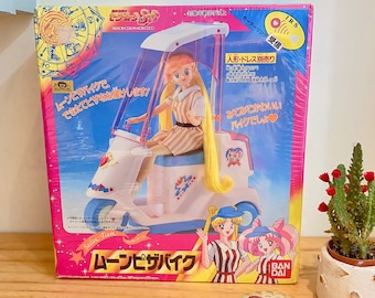 May include: A pink, blue, and white cardboard box with a picture of a toy scooter and a cartoon girl with blonde hair. The box has Japanese text that reads "Sailor Team Moon Pizza Bike" and "Ban Dai".