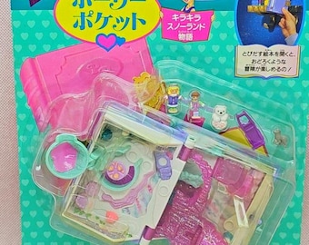 May include: A Polly Pocket Snowland Story playset in original packaging. The set includes a book-shaped case, small figurines, and accessories. The teal backing features heart patterns, Japanese text, and a graphic of a character. The clear plastic blister pack protects the contents, which include a pink structure and miniature figures.