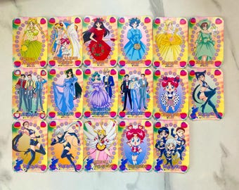 May include: A collection of seventeen rectangular Sailor Moon trading cards arranged in three rows. Each card features colorful anime-style illustrations of characters in various outfits, including formal gowns and battle uniforms, with decorative floral borders.