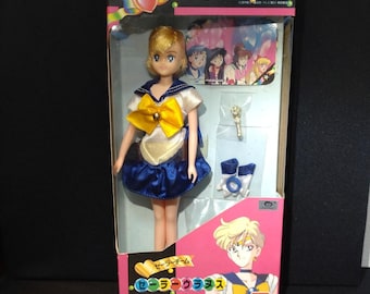 May include: A Sailor Moon S Sailor Uranus doll in original packaging. The doll has short blonde hair and wears a white and blue sailor-style uniform with a yellow bow. Accessories include a small wand and white gloves. The box features character art.