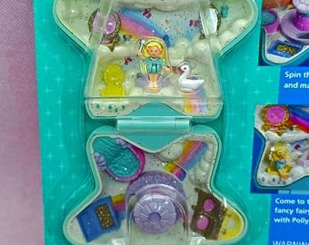 May include: A star-shaped Polly Pocket playset in teal packaging featuring a miniature blonde doll, a white swan, and a rainbow graphic. The interior includes a bed, a mirror, and a bathtub, with text on the packaging reading Spin the and make and Come to the fancy fairy with Polly and WARNING Contains small parts.