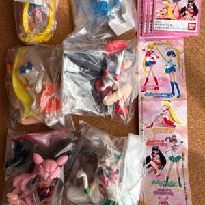 Op de afbeelding: Een verzameling Sailor Moon-personagebeeldjes in doorzichtige plastic verpakkingen, waaronder Sailor Moon, Sailor Mercury, Sailor Mars, Sailor Jupiter en Black Lady, vergezeld door een roze informatiekaart met illustraties van de personages.