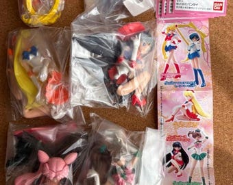 May include: A collection of Sailor Moon character figurines in clear plastic packaging, including Sailor Moon, Sailor Mercury, Sailor Mars, Sailor Jupiter, and Black Lady, accompanied by a pink informational card featuring character illustrations.