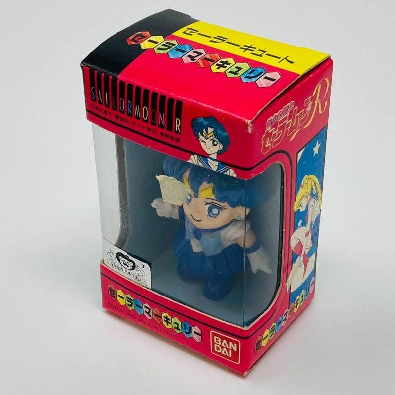 Op de afbeelding: Een Sailor Moon-figuur in een doorzichtige plastic displaydoos. De doos is rood met Japanse tekst en illustraties van Sailor Moon-personages. Het figuur is blauw en wit.
