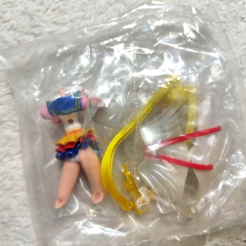 May include: A disassembled Sailor Moon action figure inside a clear plastic bag. The figure features a blue sailor collar, a multi-layered yellow and blue skirt, and flesh-toned legs. Long yellow hair pieces and white accessories are included.