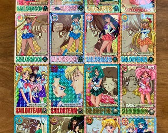 Puede incluir: Una colección de dieciséis tarjetas coleccionables rectangulares con personajes de anime de Sailor Moon. Cada tarjeta tiene un acabado holográfico prismático y muestra ilustraciones de personajes con sus nombres en inglés y japonés.