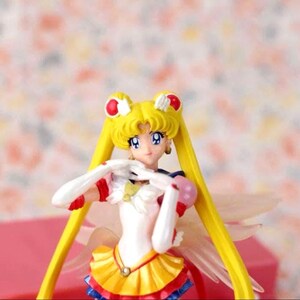 May include: A Sailor Moon figurine featuring a character with long blonde hair in twin tails, wearing a white, yellow, red, and blue outfit with gold accents. The figure has white gloves, red hair accessories, and translucent wings extending from the back.