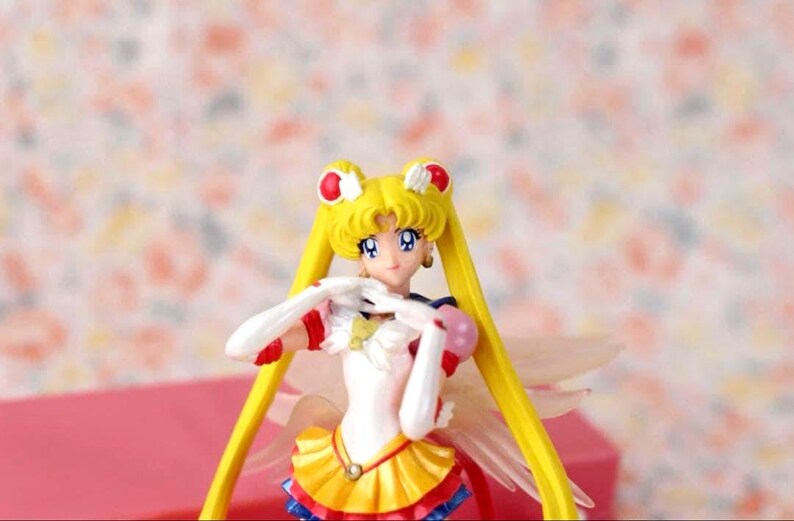 May include: A Sailor Moon figurine featuring a character with long blonde hair in twin tails, wearing a white, yellow, red, and blue outfit with gold accents. The figure has white gloves, red hair accessories, and translucent wings extending from the back.