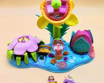 May include: A Polly Pocket playset featuring a large yellow flower structure with a rotating carousel, a pink flower-shaped house, a small bridge, and a central fountain. Two small dolls are included, one with a green skirt and one with a purple skirt.