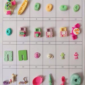 Op de afbeelding: Een verzameling kleine, kleurrijke plastic speelgoedjes, waaronder een gele kano, groene en gele vormen, miniatuurgebouwen en figuren. Het speelgoed is gerangschikt op een raster, met een verscheidenheid aan ontwerpen en kleuren.