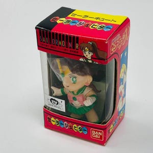 Op de afbeelding: Een Sailor Moon R Jupiter-figuur in een rode, zwarte en gele doos met Japanse tekst en een transparant plastic venster. Het figuur is groen, roze en bruin.