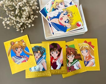 May include: A collection of Sailor Moon character trading cards featuring vibrant anime illustrations of the scouts. The cards are displayed in a white plastic storage box and spread out on a surface, showcasing gold-bordered designs and portraits.