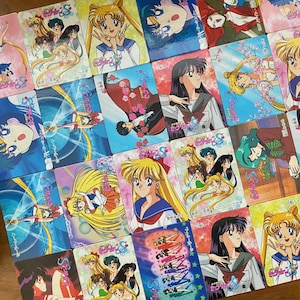 Könnte beinhalten: Eine Sammlung farbenfroher Sailor Moon Sammelkarten. Die Karten zeigen verschiedene Charaktere in Actionposen, mit lebendigen Hintergründen und japanischem Text. Die Karten sind auf einer Holzoberfläche angeordnet.