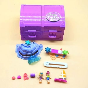 Puede incluir: Un juego de Polly Pocket morado con una caja en forma de cofre del tesoro. El juego incluye figuras en miniatura, una isla con forma de ballena, un barco y otros accesorios. La caja mide unos 10 cm de ancho.