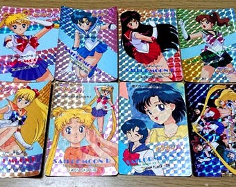 May include: A collection of eight rectangular trading cards featuring Sailor Moon characters in various poses. Each card has a holographic, prismatic finish and includes Japanese text, character illustrations, and the Sailor Moon logo.