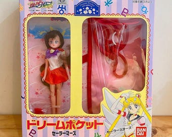May include: A boxed Sailor Mars doll from the Dream Pocket series. The box features Japanese text and illustrations of the character. The doll is wearing a red and white outfit. The box is yellow, pink, and blue.