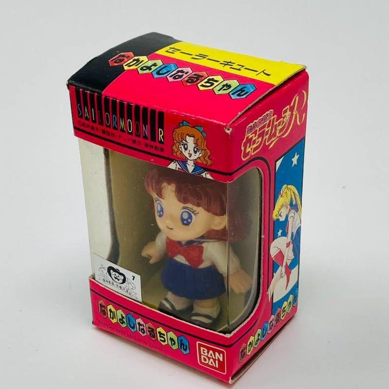 Op de afbeelding: Een Sailor Moon-figuur in een doos. De doos is rood, zwart en geel met Japanse tekst en illustraties. Het figuur draagt een blauwe rok, wit shirt en rode strik. De doos heeft een doorzichtig plastic venster.