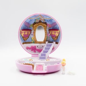 Op de afbeelding: Een roze, open miniatuurpoppenhuis met een spiegel, trap en twee decoratieve urnen. Het interieur is blauw en goud. Een klein poppenfiguur staat ernaast.