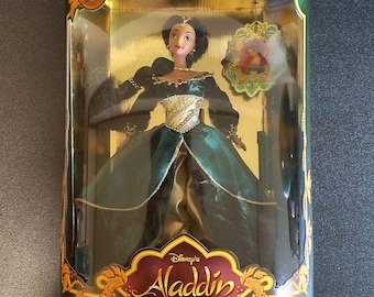 May include: A Holiday Princess Jasmine doll from the Disney Aladdin special edition series, presented in a green and gold display box. The doll wears a dark green gown with gold accents and a matching ornament is included inside the packaging.