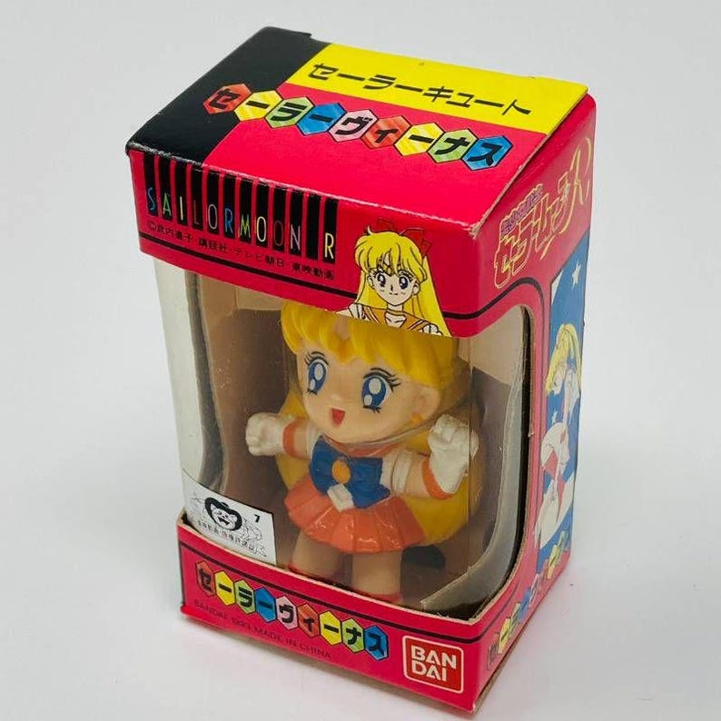 Op de afbeelding: Een Sailor Moon R-figuur in een doos. Het figuur is een stripfiguur met geel haar, een oranje rok en witte handschoenen. De doos is rood met Japanse tekst en een doorzichtig plastic venster.