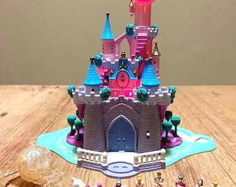 May include: A pink and gray toy castle with blue conical roofs and illuminated windows, set on a light blue base. Included are a translucent carriage with two white horses and six small character figurines in various colored outfits.