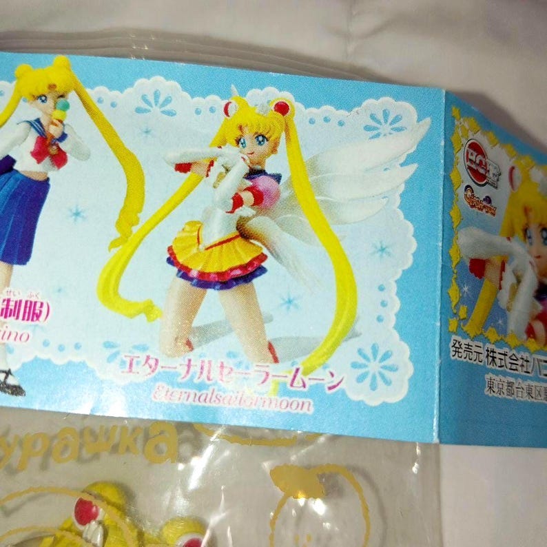May include: A light blue product packaging insert featuring illustrations of the character Sailor Moon. One image shows the character in a school uniform, while the other depicts Eternal Sailor Moon with white wings, a yellow skirt, and red accents.