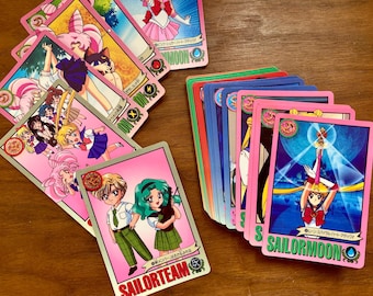 May include: A collection of Sailor Moon trading cards featuring various anime characters in colorful illustrations. The cards display pink borders, Japanese text, and character names like Sailor Team and Sailor Moon, with some cards fanned out.