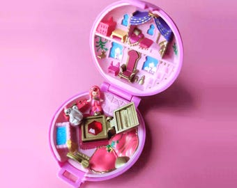 Könnte beinhalten: Ein Vintage-Miniatur-Spielset in Rosa, geöffnet, um einen detaillierten Innenraum mit winzigen Möbeln und einer kleinen Figur zu enthüllen. Das Spielset ist rund und hat einen Klappdeckel mit einem hellrosa Hintergrund.