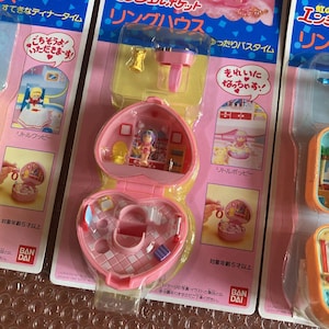 Puede incluir: Una caja de plástico rosa en forma de corazón con un anillo en su interior. La caja se abre para revelar una casa de muñecas en miniatura con una muñeca dentro. La casa de muñecas está decorada con detalles rosas y blancos. El embalaje incluye el texto "Ring House" y "Little Poppie".