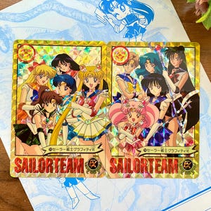 Könnte beinhalten: Zwei rechteckige Sammelkarten mit goldener holografischer Oberfläche, die Anime-Figuren zeigen. Jede Karte enthält den Text SAILORTEAM und japanische Schriftzeichen. Die Karten zeigen Gruppen von Kriegerinnen in bunten Uniformen.