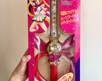 May include: A Sailor Moon Kaleido Moon Scope toy in its original pink and purple packaging. The wand features a clear, glittery shaft, a gold crown-like center, and a red handle. The box displays Sailor Moon graphics and Japanese text.