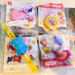 Może przedstawiać: Kolekcja zapieczętowanych zabawek McDonald's Happy Meal z motywami Polly Pocket. Zabawki obejmują różowy medalion w kształcie serca, żółty zegarek z niebieskim motylem oraz różne kompaktowe w kształcie kwiatów i pierścieni. Opakowanie zawiera japoński tekst.