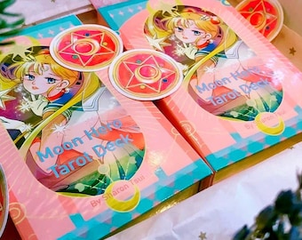 May include: Two rectangular boxes of the Moon Hero Tarot Deck by Sharon Tsui, featuring a blonde character in a sailor-style uniform. The pink and teal packaging includes circular stickers with a red star and moon emblem design.