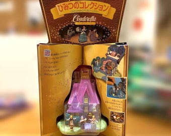 May include: A Disney Cinderella playset in a brown and gold cardboard package. The set includes a purple house with a gold roof and three small character figurines. The packaging features Japanese text, illustrations of the movie, and a clear plastic window displaying the toy house and figures inside.