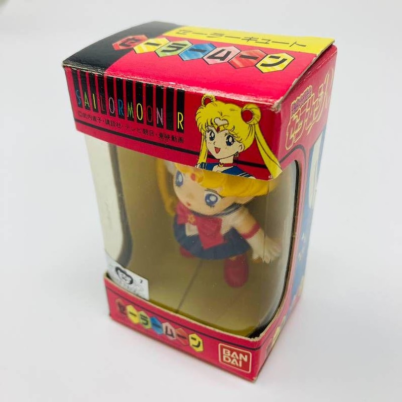 Op de afbeelding: Een Sailor Moon R-beeldje in de originele doos. De rode doos heeft Japanse tekst en een afbeelding van Sailor Moon. Het beeldje is zichtbaar door een transparant plastic venster. Het beeldje draagt een matrozenuniform.