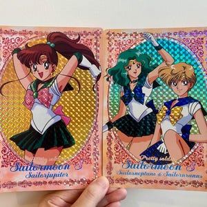 以下が含まれることがあります： セーラームーンのトレーディングカード2枚。アニメのキャラクターが描かれています。カードはホログラフィックの背景とピンクの装飾的なボーダーがあります。1枚のカードにはセーラージュピター、もう1枚にはセーラーネプチューンとセーラーウラヌスが描かれています。「セーラームーン」の文字とキャラクター名が見えます。