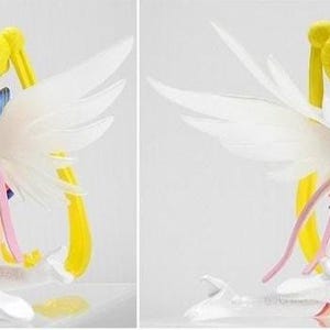 May include: A Sailor Moon figurine featuring long yellow pigtails, large translucent white wings, and a blue and white sailor-style outfit with a pleated skirt. The character is depicted in a kneeling position with white boots and pink ribbons.