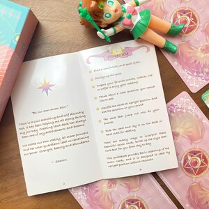 Puede incluir: Una gu&iacute;a de cartas de tarot abierta en las p&aacute;ginas dos y tres, con texto sobre princesas lunares y guardianes solares. Una peque&ntilde;a figura de estilo anime y varias cartas de tarot con dise&ntilde;os de alas rosas rodean el folleto abierto.