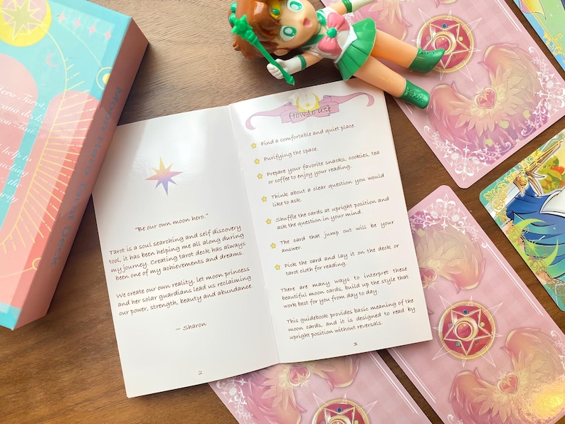 Puede incluir: Una gu&iacute;a de cartas de tarot abierta en las p&aacute;ginas dos y tres, con texto sobre princesas lunares y guardianes solares. Una peque&ntilde;a figura de estilo anime y varias cartas de tarot con dise&ntilde;os de alas rosas rodean el folleto abierto.