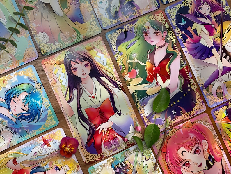 Puede incluir: Una colecci&oacute;n de cartas de tarot rectangulares con ilustraciones estilo anime de personajes con colores de pelo vibrantes, bordes de filigrana dorada y motivos simb&oacute;licos. Las cartas muestran varias figuras con atuendos detallados, incluyendo un personaje con kimono blanco y lazo rojo y otro con pelo largo verde sosteniendo una balanza.