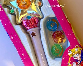 May include: A Sailor Moon Power Stick toy in its original pink packaging. The wand features a gold star head with a central orange gem, a pink crown-like base, and a light purple handle. Three additional colored gems are included in the box.