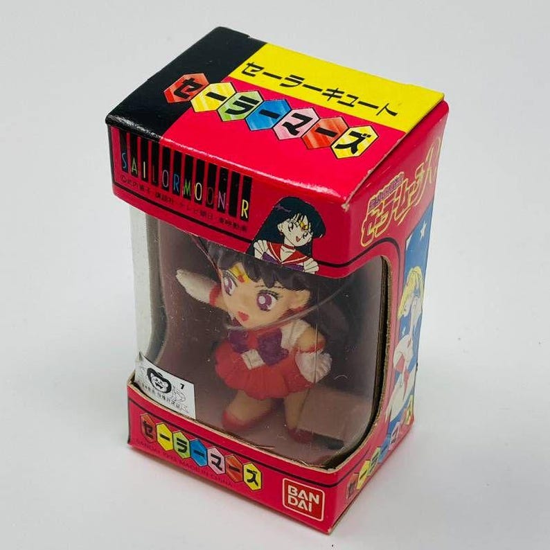 Op de afbeelding: Een Sailor Moon R-figuur in een doos. De doos is rood, zwart en geel met Japanse tekst en een doorzichtig plastic venster. Het figuur is van Sailor Mars, gekleed in een rood en wit outfit.