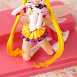 May include: A Sailor Moon anime character figurine featuring long yellow hair, a white leotard with a yellow pleated skirt, blue trim, red accents, and translucent wings. The figure is posed in a kneeling position with white gloves and boots.