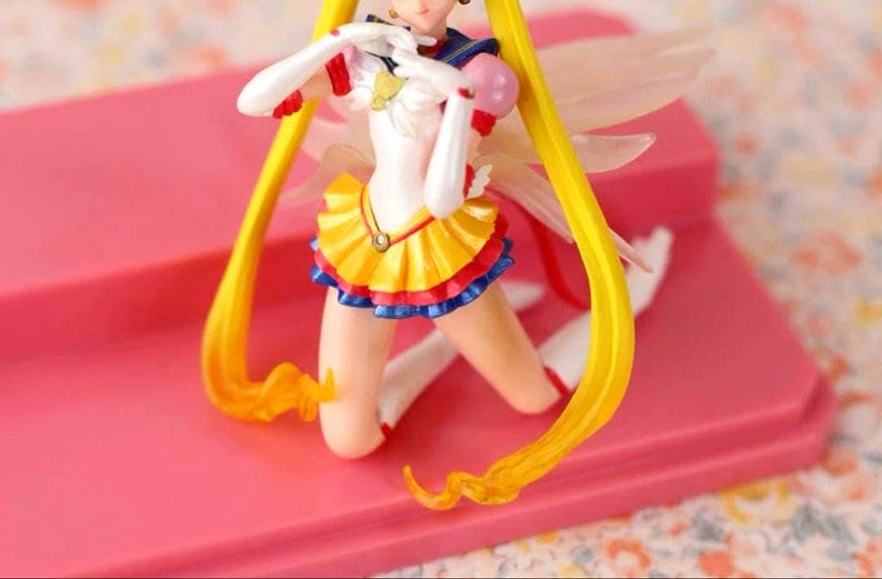 May include: A Sailor Moon anime character figurine featuring long yellow hair, a white leotard with a yellow pleated skirt, blue trim, red accents, and translucent wings. The figure is posed in a kneeling position with white gloves and boots.