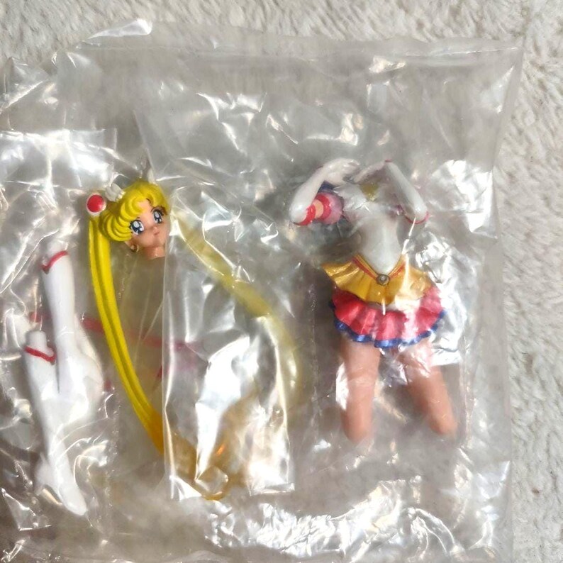 May include: A disassembled Sailor Moon anime character figurine inside a clear plastic bag. The set includes a head with long blonde hair, a torso with a white top and yellow and pink skirt, and a separate white boot, all made of painted plastic.