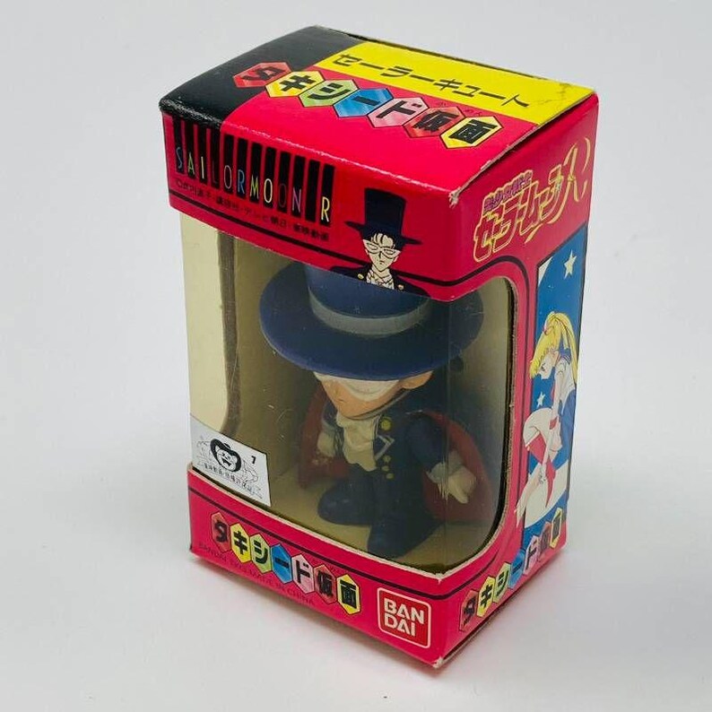 Op de afbeelding: Een Sailor Moon-figuur in een rode doos met Japanse tekst. Het figuur is van een personage met een hoge hoed en cape, zichtbaar door een transparant venster.